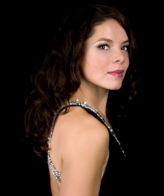 Mezzo-soprano Katija Dragojevic in an elegant black gown with silver embellishments, dark curly hair, against black background.