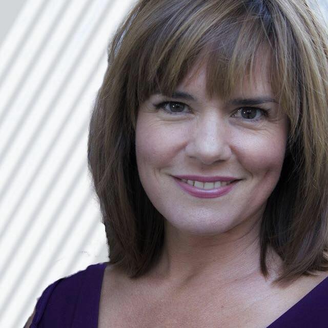 Woman with brown shoulder-length hair and bangs smiling, wearing a purple top against a striped background