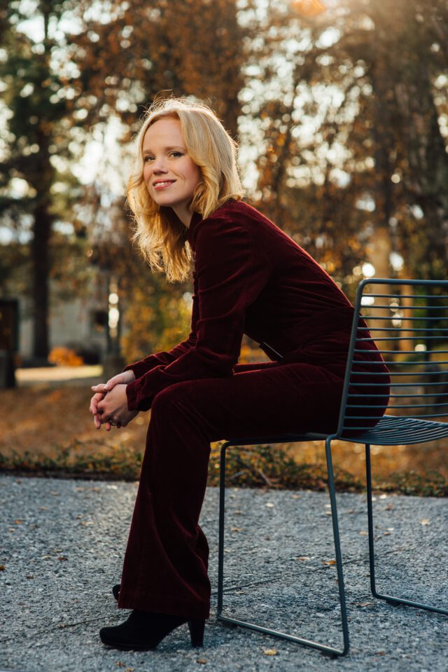 Smiling blonde woman in a burgundy velvet suit seated on a metal chair in an autumn park setting