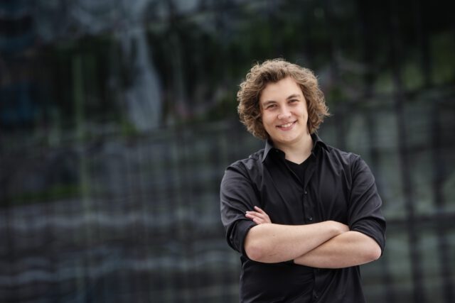 Young person with curly blonde hair smiling, arms crossed, wearing a black shirt in an outdoor setting