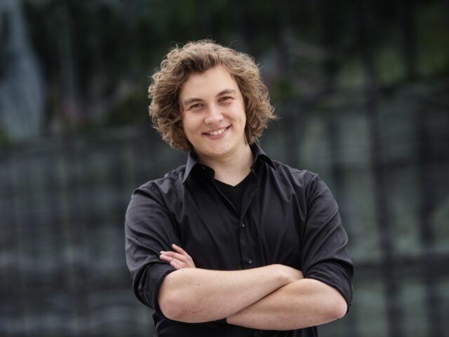 Young person with curly blonde hair smiling, arms crossed, wearing a black shirt in an outdoor setting
