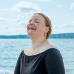 Hanne Marit Mordal Iversen laughing joyfully outdoors by a sunlit lake wearing a black top on a bright day