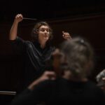 Orchestra conductor in black shirt raising baton while leading musicians in a dramatic, dimly lit concert hall performance