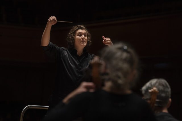 Orchestra conductor in black shirt raising baton while leading musicians in a dramatic, dimly lit concert hall performance
