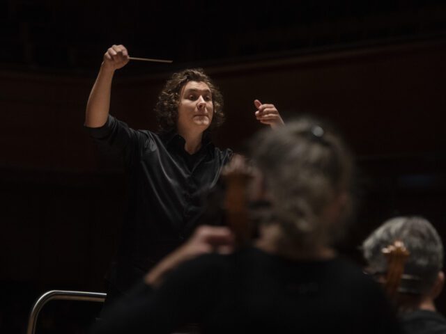 Orchestra conductor in black shirt raising baton while leading musicians in a dramatic, dimly lit concert hall performance