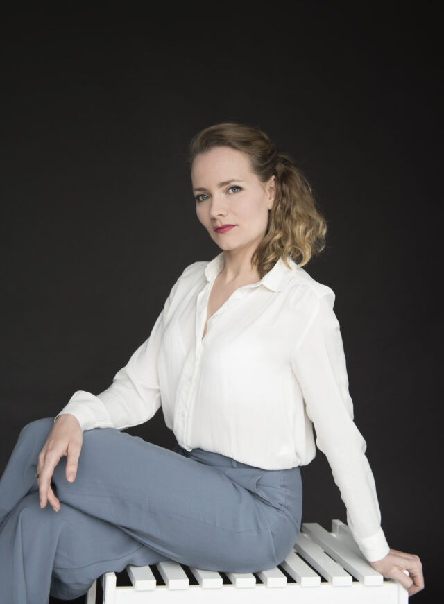 Portrait of soprano Dénise Beck seated in a white blouse and blue trousers against a dark background